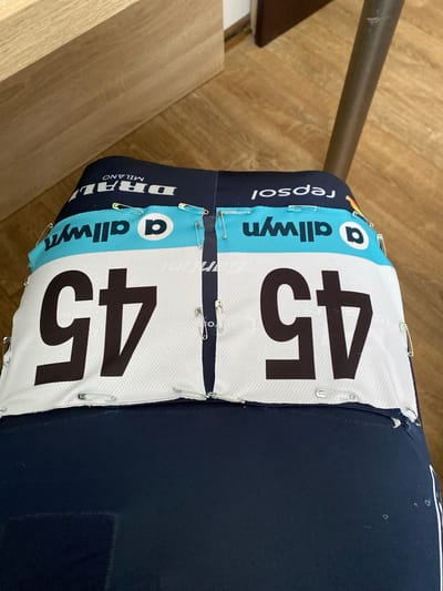 The image shows two cycling jerseys with the number "45" pinned to them with safety pins. The jerseys have the logos of "Repsol", "Allwyn", and "DRAL MILANO" on them. The jerseys appear to be pinned onto a piece of dark blue fabric, possibly a piece of furniture. The background includes a wooden floor and a metal pole. The location in the city of Bělá pod Pradědem, Czechia is not evident from the image.