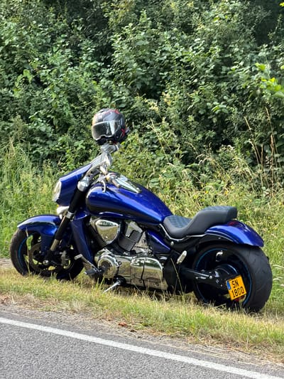 A blue motorcycle is parked on the side of a road, with its front wheel facing left and its rear wheel facing right. The motorcycle is positioned on a grassy verge with a dense green bush line behind it. A black motorcycle helmet with red accents is perched on the handlebars. The motorcycle has chrome accents on its engine and exhaust pipes. A yellow license plate with the characters "SN 1800" is attached to the rear fender. The road in front of the motorcycle is asphalt with a white line marking. The scene appears to be in a rural or semi-rural area.