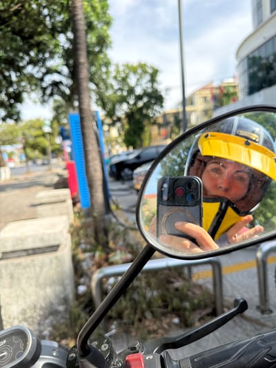 A selfie is taken in a motorcycle's rearview mirror, showing a person wearing a yellow helmet and a yellow vest, holding a smartphone. The person is in the driver's seat of a motorcycle, which is parked on a street. In the background, there are trees, buildings, and other vehicles. The scene appears to be in Đồng Hới Ward, Vietnam, as indicated by the presence of palm trees, tropical foliage, and architectural styles common in Southeast Asia. The street has a paved surface and a sidewalk. The overall atmosphere is one of urban travel and a bright, sunny day.