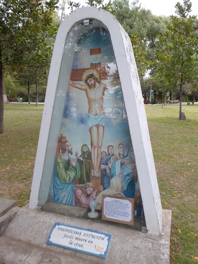 The image shows a roadside shrine in a park setting, possibly within the city of Municipio Tarija, Bolivia. The shrine is a concrete structure housing a depiction of the crucifixion of Jesus. The artwork shows Jesus on the cross, with figures mourning at the base, including Mary and several disciples. Above the cross are the letters INRI. At the base of the monument are two signs. The top sign, made of cardboard, contains a prayer in Spanish, while the bottom sign reads, "DUODECIMA ESTACIÓN" and "Jesús muere en la cruz." The shrine is located in a park with trees and grass, and benches are visible in the background.