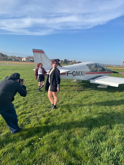 An outdoor scene captured at an aerodrome in Chonas-l'Amballan, France, depicts a photoshoot involving three individuals and a single-engine light aircraft on a sunny day.
In the foreground, a male photographer, dressed in a black long-sleeved top and dark blue trousers, is seen from behind in a slightly crouched position, holding a black DSLR camera aimed towards the subjects. The ground beneath is covered in lush green grass with scattered small white flowers.
In the mid-ground, a white Robin DR400 light aircraft, bearing the registration F-GMXI on its fuselage, is parked on the grass. It features a red stripe along its side and wings. Two female models are posing in front of and beside the aircraft. The model closer to the photographer, positioned towards the center-right, has short, curly, reddish-brown hair and wears a dark blazer over a light garment, a dark miniskirt, and black ankle boots. A tattoo is visible on her left forearm. The second model stands further back, to the left of the aircraft's tail, with shoulder-length, curly, reddish-brown hair. She wears a white turtleneck, a reddish-brown jacket, a black miniskirt, dark knee-high socks, and black ankle boots. A fourth individual with dark hair is partially visible behind the aircraft's cockpit.
The background reveals a flat, grassy expanse characteristic of an airfield, with distant residential buildings featuring red-tiled roofs and green trees under a clear blue sky dotted with scattered white clouds. Bright sunlight casts distinct shadows, indicating mid-day or late afternoon conditions.