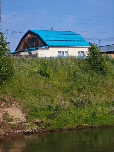 The image captures a view of a house with a bright blue, corrugated metal roof, situated on a grassy embankment overlooking a body of water. The house is painted cream with white trim around two windows and a blue picket fence runs in front of it. A street sign reading "Береговая 58" (Beregovaya 58) is visible on the side of the house, indicating its address. The embankment is covered in lush green grass and wildflowers, with a sandy, eroded slope on the left leading down to the water's edge. The water's surface is dark and reflective. In the background, power lines stretch across a clear blue sky. To the right of the main house, another building with a blue-striped roof is partially visible. The scene is consistent with a residential area in a rural or suburban setting, possibly in or near Galanino, Russia, given the street sign. There are no people or animals visible in the image.