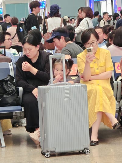 This image captures a crowd of people, likely at an airport or train station in Fengtai District, China, given the visible text "北京丰台" (Beijing Fengtai) on a blue sign in the background. The subjects are diverse, including men, women, and a young child. Many are seated, suggesting they are waiting for transportation or a flight.
The scene is busy, with people carrying luggage, including a prominent silver suitcase with wheels. One woman in a yellow outfit is holding a phone, possibly taking a selfie or a picture of the child who is peeking out from behind the suitcase. Another woman, wearing glasses, is looking at her phone. The overall mood appears to be one of waiting, with some individuals appearing tired or bored, while others are engaged with their devices.
The lighting suggests it might be daytime, but the exact time of day is not discernible. There are no clear indications of weather conditions. The setting appears to be an indoor waiting area with tiled flooring and some signage. The presence of luggage and the busy atmosphere strongly point to a travel hub.