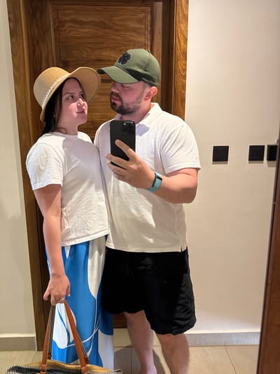 A couple takes a mirror selfie. The man wears a green baseball cap with a clover, a white t-shirt, black shorts, and a teal wristband. He holds a black smartphone. The woman wears a straw hat and a white t-shirt. A blue and white patterned beach blanket is behind her, along with a straw bag with brown straps. Three black squares are visible on the wall above them. The surrounding decor includes a wooden door frame and tan wall paneling.