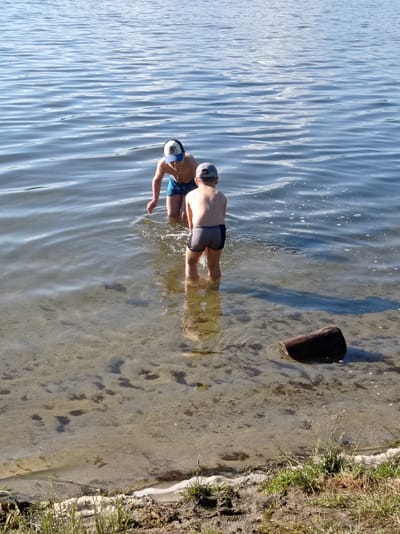 Two young boys are standing in shallow water near the shore of a lake. The boy on the left, wearing a blue and white baseball cap and blue swim trunks, is facing towards the water, with his body crouched down and his left hand extended. The boy on the right, wearing a gray baseball cap and dark swim trunks, is facing away from the viewer, with his body slightly bent. The water is clear, showing the sandy bottom and some scattered dark objects. A dark, cylindrical log rests partially submerged in the water in the foreground. The background is a vast expanse of calm, blue water under a bright sky. The scene appears to be a recreational area on the shore of a lake, possibly in or near the city of Degtyarsk, Russia, given its natural and open body of water.