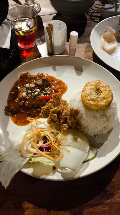 A overhead shot shows a plate of food on a wooden table. The plate contains white rice topped with a golden-brown circular fried egg. To the left of the rice is a portion of dark brown meat in a reddish-orange sauce, garnished with sliced green onions and sesame seeds. Beside the meat is a pile of crispy fried chicken pieces. A salad of shredded carrots, purple cabbage, and lettuce with cucumber slices and lime wedges completes the plate. In the background, a glass filled with a dark red beverage sits on the table next to a small white container with tongs and a small white bottle. A white plate with some light-colored crackers is partially visible to the right. The scene is dimly lit, suggesting an indoor setting, possibly a restaurant in Berastagi, Indonesia.