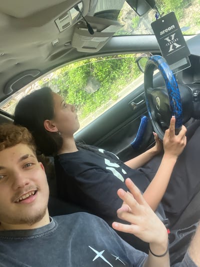 Two young individuals, a male and a female, are positioned inside a car. The male is in the foreground, looking towards the camera with a smile, his face partially visible. He is wearing a dark-colored t-shirt with a white graphic. The female is seated in the driver's seat, looking away from the camera and towards the right. She has dark hair and is wearing a black t-shirt. Her hands are on the steering wheel, which is covered with a blue patterned accessory. A black air freshener is hanging from the rearview mirror. The interior of the car, including the dashboard and door panel, is visible. Outside the car, lush green foliage suggests an outdoor environment, possibly a roadside or park. The image appears to be taken in Vlahovo, Bulgaria, based on the contextual information provided.