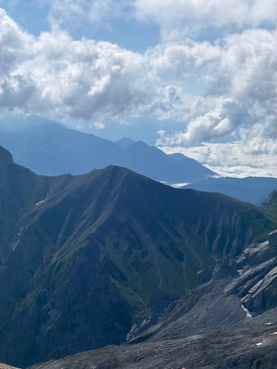 The image depicts a vast mountain landscape. In the foreground, a rugged, rocky slope dominates the lower portion of the frame, transitioning into steep, dark-colored mountains with sparse green vegetation clinging to their sides. These mountains extend upwards, their textured surfaces and deep ravines clearly visible. In the mid-ground, a prominent mountain ridge with sharp contours and shadowed valleys is the central focus. Further in the distance, multiple layers of mountains fade into a hazy blue, creating a sense of depth and atmospheric perspective. The sky above is a bright blue, filled with fluffy white clouds, some of which are illuminated by sunlight, casting subtle shadows on the mountains below. The overall scene is one of grandeur and natural beauty, characteristic of alpine environments. The location is the municipality of Wildermieming in Tyrol, Austria, a region known for its mountainous terrain and scenic vistas. There are no people or specific objects, apart from the natural landscape, visible in the image.