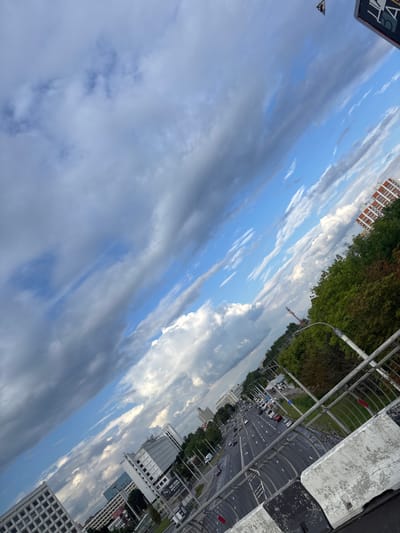 The image captures a view of a highway in the city of Senitsky Selsoviet, Belarus, from an elevated perspective, likely a bridge or overpass. The scene is characterized by a dramatic sky with a mix of dark, heavy clouds and bright, blue sky interspersed with wispy white clouds.
The highway itself is a multi-lane road with visible lane markings and traffic. Several vehicles, including cars and trucks, are in motion. The road appears to be wet, suggesting recent rainfall or humidity.
Along the sides of the highway, there are areas of green vegetation, including trees, and some urban infrastructure. Buildings of various sizes and architectural styles are visible in the background, indicative of an urban or suburban environment. One prominent building appears to be an apartment complex with orange-red brick accents. There's also a construction crane visible in the distance.
In the foreground, the edge of the elevated structure is visible, featuring a concrete barrier with black and white markings. Metal railings line the edge, consistent with a pedestrian walkway or viewing area. A signpost with some text is partially visible at the top right of the image, though the text is not clearly legible. The overall impression is that of a somewhat overcast day with dynamic cloud formations over a busy urban thoroughfare.