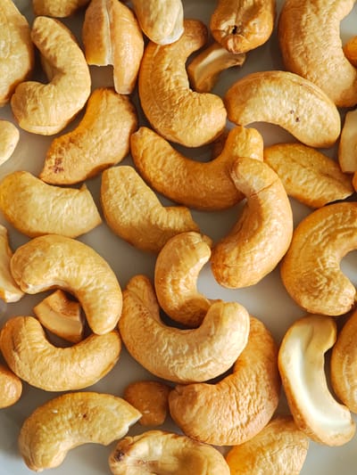 The image is a close-up, overhead shot of a pile of roasted cashew nuts. The nuts are a golden brown color and are scattered across a light grey surface. Some of the cashews are whole, while others are broken into pieces. The texture of the cashews appears slightly rough and bumpy. There are no people or animals present in the image. The lighting is even, casting minimal shadows, suggesting an indoor setting. The specific location in the city of Kursk, Russia, cannot be determined from the visual information provided.