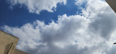 The image shows a view of the sky with clouds from an elevated perspective, likely from a rooftop in Sderot, Israel. A beige-colored building corner is visible in the lower left, with a vertical metal pipe or conduit running down its side. The sky is a vibrant blue, interspersed with white and grey cumulus clouds, suggesting a partly cloudy day. The angle of the photograph is looking upwards towards the sky.