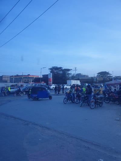 A street scene unfolds with motorcycles and auto rickshaws lining the roadside. Pedestrians gather near the vehicles. A gas station with visible signage stands beside the road. Overhead electrical wires stretch across a cloudy sky. Buildings are scattered among trees in the background. The road surface shows signs of wear.