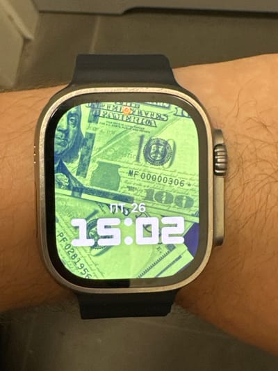 This media file shows a close-up view of a smartwatch on a person's wrist. The smartwatch face displays a background of US dollar bills. The time is shown as "20:51" and the date is "PT 26". Visible text on the watch face includes "UNITED STATES OF AMERICA", "THIS NOTE IS LEGAL TENDER FOR ALL DEBTS, PUBLIC AND PRIVATE", "JULY 4, 1976", "MF00000306", and "PF 0281956". The smartwatch has a metallic casing and a dark band. The person's wrist and arm, covered in light hair, are visible. The setting appears to be indoors, with a neutral background that is out of focus. No specific weather or location cues are apparent, other than the provided context that the location is Chociule, Poland. The image focuses on the technology and the chosen watch face.