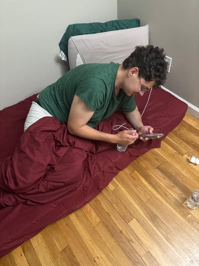 This image captures an indoor scene in Franklin Township, United States of America, depicting a person lying on their side on a blanket spread on a wooden floor. The individual, appearing to be male, has dark, curly hair and wears eyeglasses. They are dressed in a short-sleeved green t-shirt, with a glimpse of white underwear or shorts visible above a dark red blanket or comforter that covers their lower body.
The central activity involves the person using a smartphone, holding it with both hands while looking at its screen. A white charging cable is connected to the phone, draping across the blanket. Nearby, on the wooden floor, is a clear glass or plastic bottle, possibly for water, and a crumpled piece of white paper or tissue. The person's head rests on two pillows – a light grey one over a dark green one – positioned against a plain light-colored wall. The floor consists of light-toned wooden planks. The scene suggests a relaxed, casual indoor setting, likely a bedroom or living area. No specific time of day is discernible, and no explicit text is clearly visible on any objects.