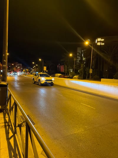A night-time photograph captures a street scene in Ibiza, Spain. A white taxi is in the foreground, turning right. The street is wet, reflecting the yellow streetlights. A concrete barrier, illuminated from above by a bright light, runs along the right side of the road. In the background, the neon sign of "Ibiza Gran Hotel" is visible, along with palm trees and streetlights. Other cars are present further down the road, and a street pole with a trash can is on the left side of the frame.