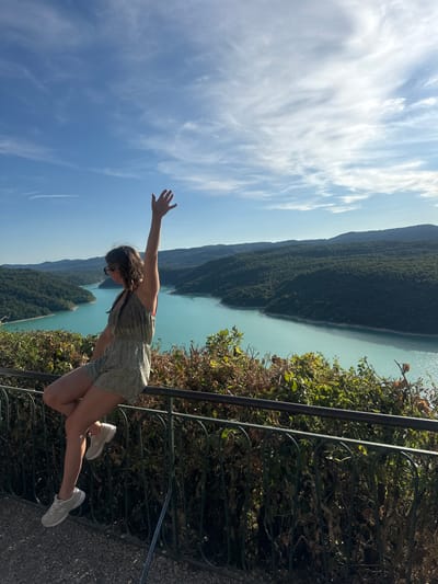 In Moirans-en-Montagne, France, a young woman is captured in a full-body, eye-level shot, perched on a black metal railing. She is facing left, with her right arm raised in a waving gesture. The woman wears a patterned romper and white sneakers. Behind her, a vast expanse of turquoise water, likely a lake or reservoir, is nestled between lush green, tree-covered hills under a bright blue sky with scattered white clouds. The scene is framed by the railing in the foreground, with dense green foliage growing behind and over it.