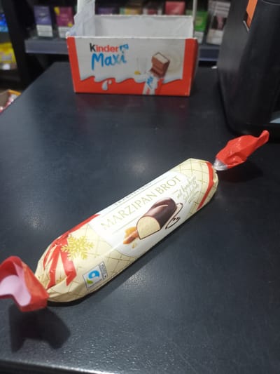The image shows a close-up view of a candy bar with a festive, Christmas-themed wrapper lying on a dark, textured surface. The candy bar is elongated and cylindrical, with red foil wrappers twisted at both ends, one of which is partially open, revealing a light pink interior. The main wrapper is predominantly cream-colored with a delicate grid pattern. It features festive red ribbons and a gold snowflake, suggesting a holiday theme. Prominently displayed on the wrapper is the text "MARZIPAN BROT" in a golden font, with smaller text below it indicating "mit knackiger Schokolade," which translates to "with crunchy chocolate."

To the left of the candy bar, partially visible, is a small, red and white cardboard box. The box has "Kinder" and "Maxi" printed on it in bold blue and black lettering, respectively. This box suggests that the candy bar may be part of a larger Kinder product offering or a display for them. In the background, out of focus, are shelves stocked with various products, some of which appear to be cigarette packs. The lighting suggests an indoor setting, likely a store or checkout counter, given the presence of what appears to be a cash register on the right side of the frame. The overall impression is of a retail environment, with a focus on the individually wrapped marzipan candy. The text "FAIRTRADE" and a circular logo are also visible on the candy bar wrapper, indicating ethical sourcing of cocoa. The location context provided is Bilhorod-Dnistrovskyi, Ukraine.