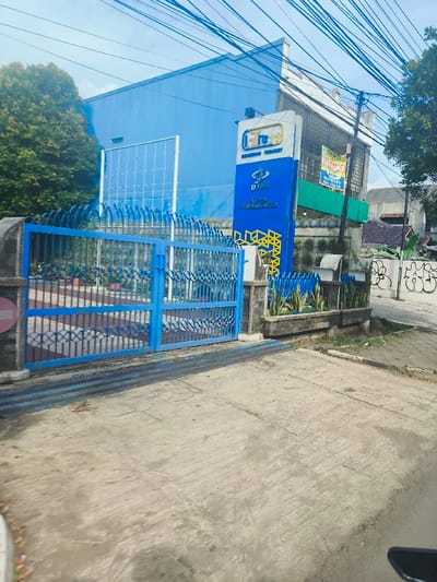 The image shows a blue gate with a blue wall behind it. The gate is made of metal bars and is closed. There is a sign on the wall that reads "Fre" in large letters. There are several electrical wires overhead, and the ground is covered in a layer of dirt and debris. The image was taken in Purwakarta, Indonesia at coordinates -6.54 latitude and 107.441 longitude. There are no people visible in the image.