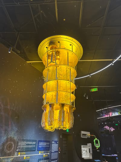 A large, golden-hued dilution refrigerator, a component integral to quantum computing, hangs centrally suspended from a dark, mesh-gridded ceiling in an exhibition space located in Guangming District, China. The apparatus is multi-tiered, featuring stacked cylindrical platforms connected by numerous vertical conduits and a dense cluster of yellow and multi-colored wiring at its lowest point. The room's walls are dark. The left wall features display panels with text in Chinese, showcasing a timeline with visible years including 2019, 2020, 2021, and 2023, set against a background depicting a starry, cosmic pattern. The right wall presents further technological exhibits, including a glowing neon green tube and abstract illuminated designs, along with a green exit sign displaying a left-pointing arrow. Overhead track lighting illuminates the exhibits. No individuals are distinctly visible.