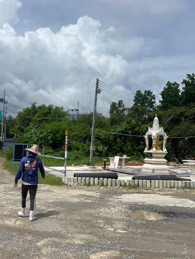 The image depicts a person walking away from the camera on a gravel road in Pa Khlok, Thailand. The person is wearing a blue long-sleeved shirt, dark pants with white stripes, a wide-brimmed hat, and white boots. They are carrying a brown bag slung over their shoulder. To the right of the person, there is a small decorative shrine with white and gold accents, which appears to be in a newly constructed or renovated area. The area around the shrine is paved with stone tiles and bordered by a low fence made of concrete posts and dark cylindrical objects. Beyond this area, lush green trees form a dense background. In the foreground, the gravel road has several puddles of water, indicating recent rain. Utility poles and power lines are visible against a cloudy sky. A blue sign board is present to the left of the frame. The overall scene suggests an outdoor location that may be a public space or a construction site undergoing development.