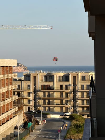 The image depicts a coastal scene in Ivanovići, Montenegro, under a clear sky. In the foreground, a partially constructed building with exposed brickwork and concrete structures is visible on the left, suggesting ongoing development. To the right, a modern apartment building with beige facades and multiple balconies dominates the mid-ground. Several air conditioning units are mounted on the exterior walls of this building.
A paved road curves through the scene, with a few individuals walking on the sidewalk. A white SUV is parked on the side of the road near the apartment building, and a black car is parked further down the road.
In the background, the Adriatic Sea stretches towards the horizon, with a small, rocky island visible to the left. A colorful parasailer is in the air over the sea, adding a recreational element to the scene. A long, white construction crane arm extends across the upper portion of the image, indicating further construction activity in the area. The overall impression is one of a developing coastal resort town.
