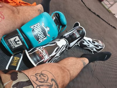 The image captures a first-person perspective of a person resting on a textured, dark mat floor, likely in a combat sports training facility. The person's legs, which are hairy and adorned with several tattoos, are prominent in the foreground. One leg wears camouflage shorts and a large tribal-style tattoo on the thigh. The other leg is partially covered by orange fabric, possibly shorts or a towel, and has a tattoo on the ankle area that reads "BELIEVE". Both feet wear black ankle-length socks, one of which has "KIPRUN" visible on the sole/top.
Next to the person are pieces of combat sports equipment. A pair of teal and black boxing gloves, branded "BUDDHA PROFESSIONAL PREMIUM FIGHT WEAR," is positioned near one leg. Beside them, a pair of black and white shin guards, featuring a flame design and branded "IMPACTO," rests on the mat. A small tag with "IMPACTO" and "www.impactoBoxing.com" is also visible. The overall scene suggests a break during or after a training session for boxing, Muay Thai, or kickboxing. The setting is consistent with an indoor gym in an industrial area, such as Polígon Industrial De Picassent, Spain.
