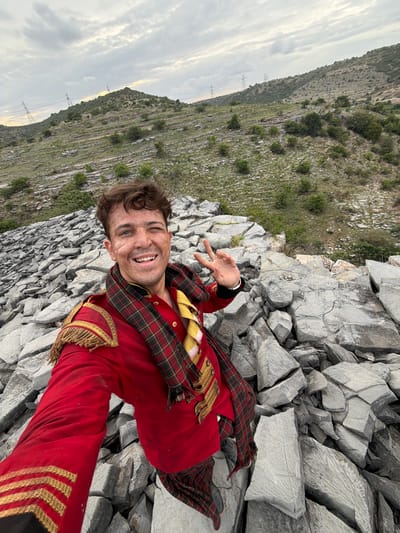 A young man in a striking red uniform adorned with gold trim and a tartan scarf smiles for a selfie taken outdoors. He is situated amidst a rocky, mountainous terrain with lush green vegetation climbing the slopes of a hill in the background. The sky is overcast with diffused light, suggesting it may be early morning or late afternoon. The man's facial expression is joyful and energetic, and he playfully holds up two fingers in a peace sign. The rocky ground he is sitting on appears to be a landslide or quarry with large, broken stone fragments. The overall scene suggests an adventurous or dramatic setting, perhaps a filming location or a location of personal significance. The uniform could indicate a costume for a play, performance, or historical reenactment. The man's freckled skin and tousled hair add to his natural and approachable demeanor. There is no visible text in the image.