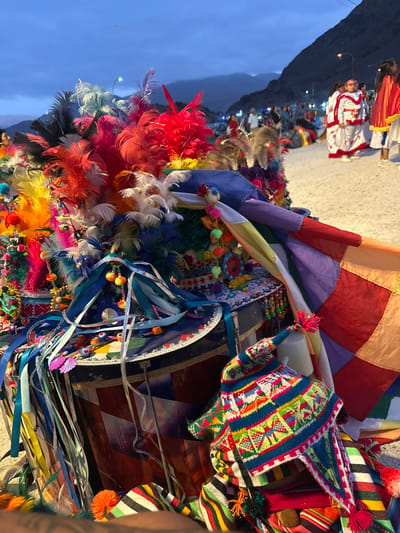 Vibrant, colorful feathers burst from a bundle, adorned with beads and ribbons. Adjacent, a geometric-patterned hat rests on a drum decorated with streamers and what appears to be a political symbol. A patchwork flag or cloth with various colors and squares sits atop the arrangement. In the background, figures in traditional clothing stand on what looks like a stage or street, set against a mountainous backdrop under a dusk sky.