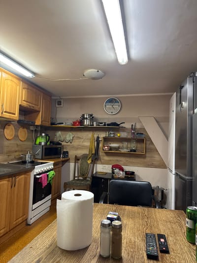 The image depicts a rustic kitchen in Wąwelnica, Poland. The kitchen is equipped with wooden cabinets, a white oven with colorful towels draped over it, and a microwave. Above the counter, shelves hold various kitchenware, including pots, pans, and a blender. A round clock hangs on the wall, and a tall refrigerator stands to the right. In the foreground, a wooden table is set with a roll of paper towels, two shakers, and two remote controls. A can of beer is also visible on the table. The lighting appears to be from fluorescent ceiling fixtures. The overall impression is one of a lived-in, functional kitchen.