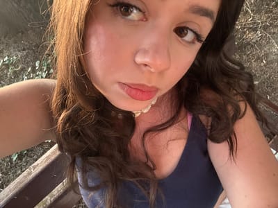 A young woman with long, wavy brown hair and brown eyes takes a selfie outdoors, possibly in the city of Garmen, Bulgaria. She is wearing a dark blue tank top with pink straps visible underneath. The background appears to be a natural setting with dry vegetation and wood, suggesting she is sitting on a wooden bench. The lighting indicates it may be late afternoon or early evening, with warm sunlight illuminating her face and hair.
