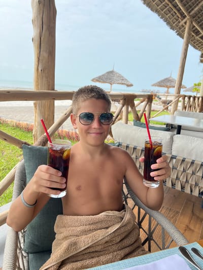 A young male is seated on a shaded outdoor patio overlooking a beach and ocean on the island of Zanzibar, Tanzania. The individual, appearing to be an adolescent, has short, light brown hair and wears aviator-style sunglasses with dark lenses. He is shirtless, with a light brown towel wrapped around his lower body. He holds a tall glass in each hand, both containing a dark beverage, ice, a lemon slice, and a red straw, extending them forward with a smile directed towards the camera. He occupies a grey-colored chair with a woven back and a teal cushion. A wooden table with a light blue placemat and silverware is partially visible in the foreground. The patio structure features rustic wooden posts and railings, supporting a thatched roof. Beyond the railing, a strip of green vegetation borders a sandy beach. The ocean, characterized by calm, light blue water, extends under a light blue sky. Several thatched beach umbrellas are present on the sand in the mid-ground.