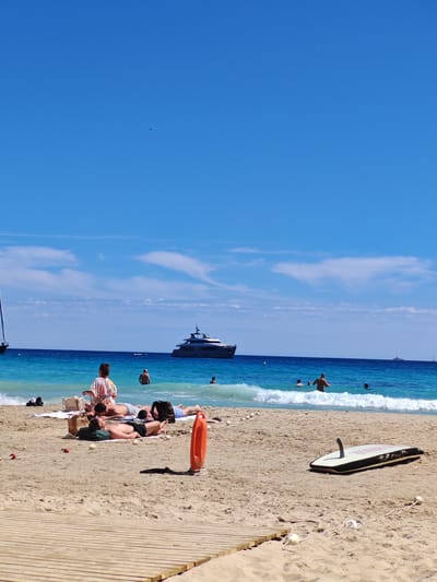 The image depicts a beach scene in Sant Agustí, Spain on a sunny day. People are sunbathing and swimming in the turquoise water. A large yacht sits in the background on the water. The beach contains a wooden boardwalk at the bottom, a lifeguard floatation device and a surfboard sitting on the sand. An airplane is also visible in the upper part of the sky.