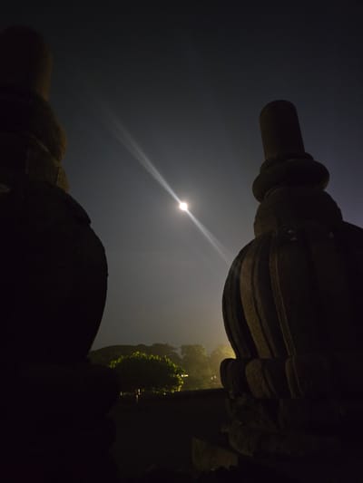 The image, taken at night, shows two ornate stone structures framing a view of the sky. The structures appear to be components of a building or monument. The sky is dark with a bright light source, likely the moon, positioned slightly left of center, with a visible beam of light. Beyond the stone structures, on the right, is a partially obscured area with trees or foliage that are lit up in a very light yellow color. The location is Bokoharjo, Indonesia.