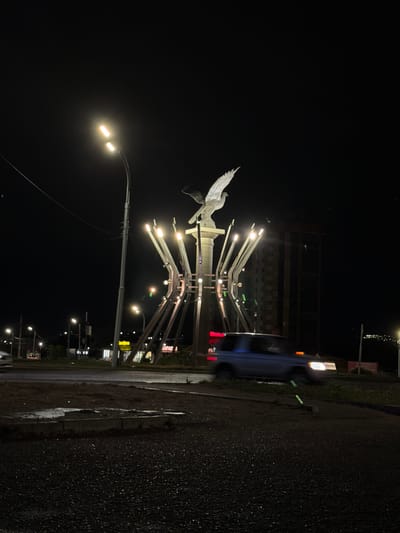 The image depicts a monument featuring two eagles at night in Ulan-Ude, Russia. The monument is illuminated by spotlights and is situated in a roundabout. A street lamp with two bulbs is visible to the left of the monument. In the background, a tall building and lights of other buildings can be seen. A blue SUV is driving through the roundabout, its headlights on, and motion blur indicates movement. The ground is paved with asphalt and concrete, with some wet patches suggesting recent rain. The sky is dark, indicating it is nighttime.