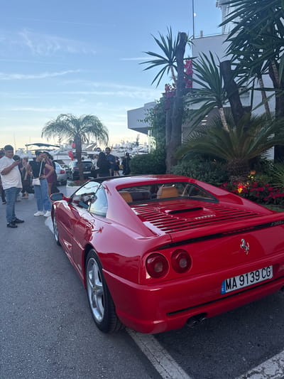 The image displays a vibrant red Ferrari F355 GTS parked on a street in Nueva Andalucía, Spain. The car, with its iconic prancing horse emblem, is the primary focus. In the background, a row of yachts suggests the setting is near a marina or port. Palm trees and lush greenery frame the scene, indicating a warm climate.
Several people are gathered around the car, observing it. Their casual attire and relaxed stances suggest a leisurely atmosphere. Some are taking pictures, while others are engaged in conversation. The overall mood appears to be one of admiration and enjoyment, characteristic of a car enthusiast gathering or a public event.
The time of day seems to be late afternoon or early evening, with the sky displaying a soft, diffused light and a few wispy clouds. The weather appears pleasant, conducive to outdoor activities. A license plate with the inscription "MA 9139 CG" is clearly visible on the rear of the Ferrari. The street is paved with asphalt and bordered by a sidewalk. The overall scene conveys a sense of luxury and an appreciation for fine automobiles within a beautiful Mediterranean setting.