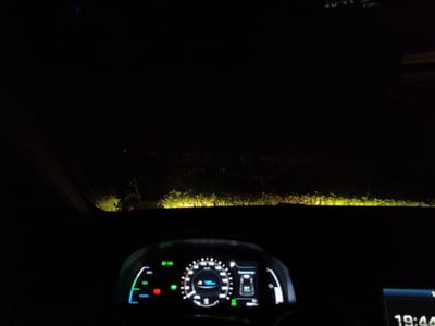 The image presents a night-time perspective from inside a vehicle, looking forward through the windshield in Villa di Gemmano, Italy. The lower portion of the frame displays a digital instrument cluster and dashboard elements. The instrument cluster shows a speed reading of "100" and "106km," along with illuminated indicators including a green "ECO" mode, a red battery/charging symbol, a green arrow indicator, and a blue warning light. An auxiliary display to the right shows the time as "19:44."
Outside the vehicle, the immediate foreground is illuminated by the vehicle's headlights, revealing bright yellow-green patches of vegetation, consistent with bushes or tall grass, directly in front and to the sides. Beyond this headlight-illuminated area, the scene is dark, with distant, faint scattered lights visible across the horizon, indicating a remote settlement or infrastructure. The sky above is entirely dark. No individuals are visible within the frame. The scene suggests a stationary or slowly moving vehicle at night in a rural or semi-rural elevated location.