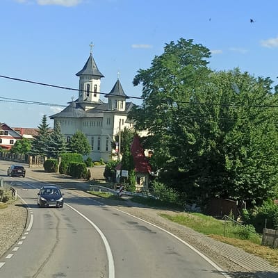 The image depicts a view of a street in Milișăuți, Romania, with a prominent church and lush greenery. A black Peugeot station wagon is driving on the road in the foreground, followed by another vehicle further back. The church, with its distinctive tall steeples topped with crosses, is situated to the left of a large, mature tree. Adjacent to the church, there are other structures and various types of trees and shrubs. The sky is blue with a few scattered clouds. A utility pole and wires are visible in the upper portion of the image. The road is paved and has white lane markings.