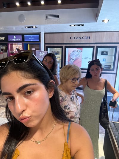 Four females are depicted inside a luxury retail establishment, identifiable by prominent "HENNESSY," "COURVOISIER," "MARTELL," and "COACH" branding on illuminated display cases. This environment suggests a duty-free store or luxury goods retailer, consistent with locations such as the Cozumel International Airport or a cruise port shopping area in San Miguel de Cozumel, Mexico.
In the immediate foreground, a young adult female with long dark hair, wearing a mustard yellow spaghetti strap top and a gold layered necklace, is capturing a selfie. Her lips are slightly pursed, and dark sunglasses rest on top of her head as she looks directly into the camera. Behind her, a second young adult female with dark hair is partially visible. To the center-right, an elderly female with short blonde hair and dark-rimmed glasses, dressed in a floral short-sleeved top, looks downwards and to her left. To the far right, a third young adult female with long dark hair, also wearing sunglasses on her head, stands in a light green polka-dot midi-dress. She carries a dark tote bag on her left shoulder and holds a black smartphone in her right hand.
The store's background features multiple display units. On the left, illuminated shelves showcase bottles of Hennessy, Courvoisier, and Martell. On the right, a prominent "COACH" sign is situated above framed displays, one featuring a large image of a wristwatch and another holding smaller Coach-branded items. A "Maui Jim" eyewear display is also partially visible. The interior is brightly lit by recessed ceiling lights and features a light-colored, reflective floor.
