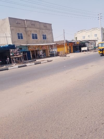 The image depicts a daytime street scene in Chiranchi, Nigeria, characterized by commercial activity. In the foreground, an asphalt road, partially covered in light-colored dust or sand, extends from the lower left to the upper right. A concrete curb delineates the road from a dirt shoulder where several single-story commercial structures are located.
On the left, a multi-story building, appearing unfinished or unpainted concrete, features three rectangular windows with blue frames on its second floor. Below this building, a row of shops with varied facades and open fronts line the street. The leftmost visible shop has a stack of blue pipes next to it. Two individuals, one standing in a light-colored shirt and another partially obscured, are visible near these shops on the left. A wooden stand displays items in front of one shop. Further right along the shop line, a dark-colored motorcycle is parked facing right. Signage with partially legible text, including "SPARE PARTS," is present on some shop awnings.
In the mid-ground right, a light-yellow three-wheeled vehicle, possibly a Keke Napep, is partially visible on the road, oriented towards the upper right, suggesting movement away from the viewer. In the background on the right, another multi-story building, light-colored and featuring multiple smaller windows, stands under a clear, bright sky. Overhead utility lines are visible across the upper portion of the image. The overall environment suggests a bustling, dry, urban commercial area.
