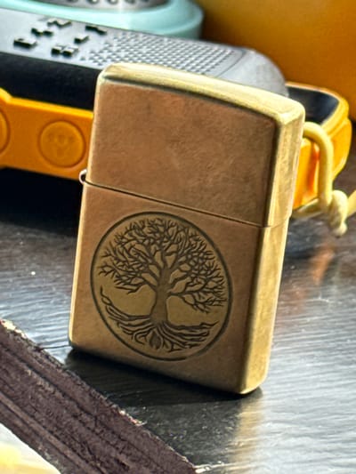 The image features a close-up, overhead view of a brass Zippo lighter with an engraved tree of life design. The lighter is resting on a dark, textured surface, possibly a wooden table. To the left of the lighter, a portion of a light blue item with a textured pattern is visible, and behind it, a bright orange object with circular indentations can be seen. A thin, light-colored cord is attached to the lighter, looping over the orange object. The lighting suggests an indoor setting. No people or specific time of day or weather cues are apparent. The image is focused on the lighter and its immediate surroundings, with the background elements blurred.