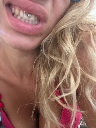 This is a close-up, horizontally oriented, low-angle shot of a person's face and upper chest. The subject appears to be a woman with blonde hair, which is messy and covers a portion of her face and neck. Her mouth is open, revealing her teeth and a small hoop piercing on her lower front teeth. Her lips are pink and appear slightly glossy. She is wearing a pink garment, possibly a bikini top or a dress, with some dark floral pattern visible. The setting appears to be outdoors, with natural light illuminating the scene. The overall mood is casual and unposed. There is no visible text in the image.