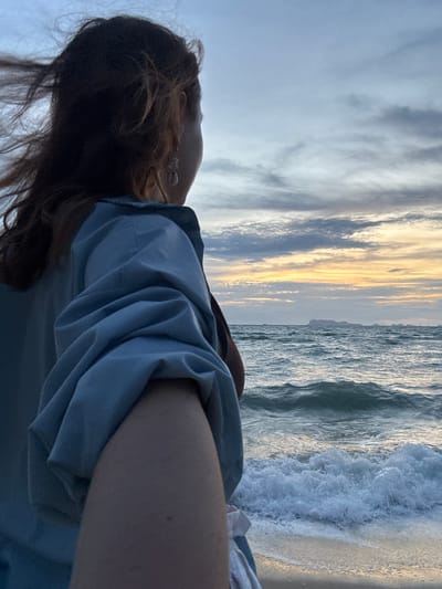 The image shows a person's arm and head from a side-view, looking out towards a beach. The person is wearing a light blue button-up shirt and has long, dark hair. They are wearing earrings. The scene is set at the beach during sunset, as indicated by the warm, golden hues in the sky and the gentle waves crashing on the shore. The sky is filled with dramatic clouds, some appearing dark and others illuminated by the setting sun. The ocean water is a deep blue, reflecting the colors of the sky. The mood appears to be peaceful and contemplative, with the individual seemingly lost in thought while observing the natural beauty around them. There is no visible text in the image.