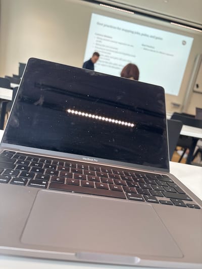 Here is a detailed description of the image:
**Scene Overview**
The image presents a close-up, slightly angled view of a MacBook Pro laptop. The focus is primarily on the laptop's screen and keyboard. The background shows a lecture-style setting, with a projection screen visible.
**Objects**
* **MacBook Pro:** The laptop is the most prominent object in the frame. The display screen is partially closed, showing reflections and dust particles. The keyboard is visible, with keys bearing standard alphanumeric and control symbols. The laptop's trackpad is also visible to the right of the keyboard.
* **Projection Screen:** In the background, a projection screen displays what appears to be a presentation. Parts of the content are visible.
* **Chairs:** Below the laptop, dark chairs can be seen.
**People**
* **Partial Figure:** On the projection screen, there are partial images of individuals. One person is visible in the lower center of the screen, wearing what appears to be a dark shirt.
**Actions and Interactions**
* The laptop is placed on what seems to be a surface (perhaps a desk or table), indicating it is either in use or in standby.
**Environment**
* **Indoor Setting:** The setting is clearly indoors, likely in a classroom or conference room.
* **Lighting:** The lighting is somewhat bright, which contributes to the reflections on the laptop screen.
**Location Information**
Based on available information, there's nothing in the image to verify this is Krems an der Donau.