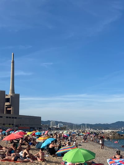 This is a daytime, wide-angle, vertical shot of a crowded beach with a prominent industrial structure in the background, likely the Besòs thermal power station in Sant Adrià de Besòs, Spain. The sky is clear blue with a few wispy clouds. The beach is filled with people sunbathing, sitting under colorful umbrellas, and walking. In the foreground, people are relaxing on towels and in beach chairs. The sea is visible in the middle ground, with some people swimming. Beyond the beach, a rocky breakwater separates the sea from a harbor with sailboats and other vessels. Further in the background, a city skyline with buildings and hills can be seen. The overall scene depicts a lively beach day with a juxtaposition of leisure activities and industrial architecture.