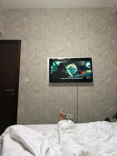 This image captures an indoor scene within a bedroom in Business Bay, United Arab Emirates. The perspective is from the bed, looking towards a wall-mounted television and a closed door.

The lower half of the frame is dominated by a white bed, covered with a white duvet and sheets. A bare human foot, belonging to an unseen individual lying in the bed, extends from beneath the duvet on the right side.

The wall directly facing the bed features a distinctive wallpaper with a light, marbled pattern, characterized by swirls of off-white and pale grey, accented with scattered flecks and streaks of gold or a similar metallic finish.

A flat-screen television is mounted centrally on this wall, enclosed within a white frame or recess. The TV is active, displaying a scene from what appears to be a film or television show, featuring a dark, complex interface with multiple data overlays and a silhouetted figure. English subtitles are visible at the bottom of the screen, stating: "Good Lord. We got seven more bogeys approaching." A black cable descends vertically from the TV, leading towards two white electrical wall sockets located below it, to the right of the visible foot.

To the left of the television, a dark brown door, possibly made of wood, is partially visible, equipped with a horizontal metallic handle. Between the door and the TV, on the marbled wall, a square white light switch is present. The ceiling, visible at the top of the image, is plain and light-colored. On the far right, next to the bed and partially obscured, a dark object, possibly footwear, rests on the floor.