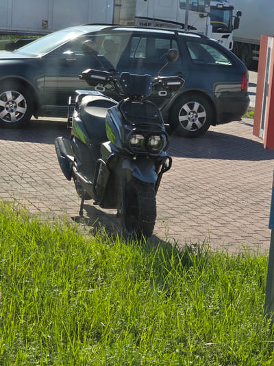 The image shows a dark colored car parked next to a motorcycle on a brick-paved surface. The motorcycle is parked in front of the car. The scene is outdoors with a large area of green grass to the right of the paved surface. A large white truck is visible behind the car, with the text "DAF XF EDITION" clearly visible on its side. The sunlight suggests it is daytime, and the shadows indicate the sun is relatively high in the sky, likely late morning or early afternoon. The overall impression is of vehicles parked in an industrial or commercial area. There are no people visible in the image. The location is Pruszcz Gdański, Poland.