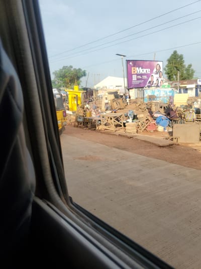 The image depicts a street scene in Goni Gara, Nigeria. In the foreground, the dark interior of a vehicle is visible, with the edge of a window frame and a black fabric lining the left side of the frame. The road surface in front of the vehicle is paved concrete.
Beyond the road, the scene opens up to a bustling market or roadside trading area. A yellow kiosk or small building is prominent on the left, with a portion of a yellow motorized tricycle parked nearby. Various stalls and makeshift structures made of wood and corrugated metal are set up along the roadside, displaying goods and seating areas. Several wooden chairs and tables are stacked and arranged.
People are visible in the market area, some appearing to be vendors or shoppers. One woman in a colorful patterned dress stands near a stall on the right. Other individuals are engaged in activities related to the stalls, such as arranging items or sitting. A large billboard is situated in the background, advertising "BMore" with images of women. The environment appears to be outdoors, with trees and some buildings in the distance, under a hazy sky. Power lines are visible stretching across the upper portion of the image. The overall impression is one of a vibrant, informal commercial district.
