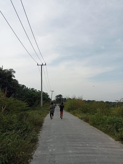 The image depicts a paved road with vegetation lining both sides, stretching into the distance under a cloudy sky. In the foreground, two individuals are seen running away from the camera on the road. The individual on the left wears a light-colored long-sleeved shirt and dark pants, while the person on the right wears a dark t-shirt and orange pants. Several utility poles with power lines are visible along the left side of the road. The environment appears to be rural, with lush greenery and some distant trees. The scene is captured during daylight hours, likely late afternoon or early morning given the overcast conditions. The image does not provide specific landmarks to pinpoint the exact location within Teluk Kenidai, Indonesia, but suggests a semi-urban or rural outskirts area characterized by residential or utility infrastructure and abundant natural vegetation.
