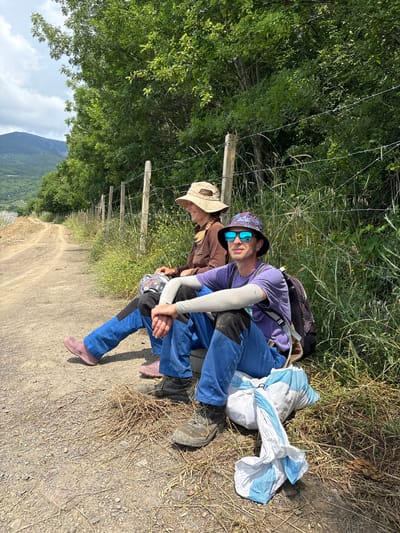 Two individuals are seated on the ground next to a dirt path and a barbed wire fence. The person on the left is wearing a brown long-sleeved shirt, a straw hat, and pink boots. They are holding a patterned item in their lap. The person on the right is wearing a purple t-shirt, a patterned bucket hat, sunglasses, and blue pants with black patches. They have a backpack on and are wearing arm sleeves. In front of them are a white and blue bag and some dried grass. The background shows a tree line, a dirt road, and a hilly landscape under a cloudy sky. The scene appears to be outdoors in a rural or natural setting.