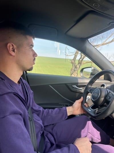 The scene depicts the interior of an Audi vehicle, specifically from the driver's perspective, located in Kozar Belene, Bulgaria. A male individual is seated in the driver's seat, shown in profile, looking forward through the windshield. He has short, dark hair with a fade haircut and a short goatee. He is wearing a purple tracksuit, consisting of a zip-up jacket and matching pants, and is secured by a black seatbelt across his chest and lap. His right hand rests on the upper-left quadrant of a black, flat-bottomed steering wheel bearing an Audi logo and an "S line" emblem. The vehicle's interior features dark dashboard and door paneling, with a silver-colored door handle visible. Outside the vehicle's side window, a clear blue sky is visible above a vast, bright green field, indicative of agricultural land. A single, bare-branched tree stands outside, partially reflected in the vehicle's side mirror. A small, reflective object is suspended from the car's overhead console.